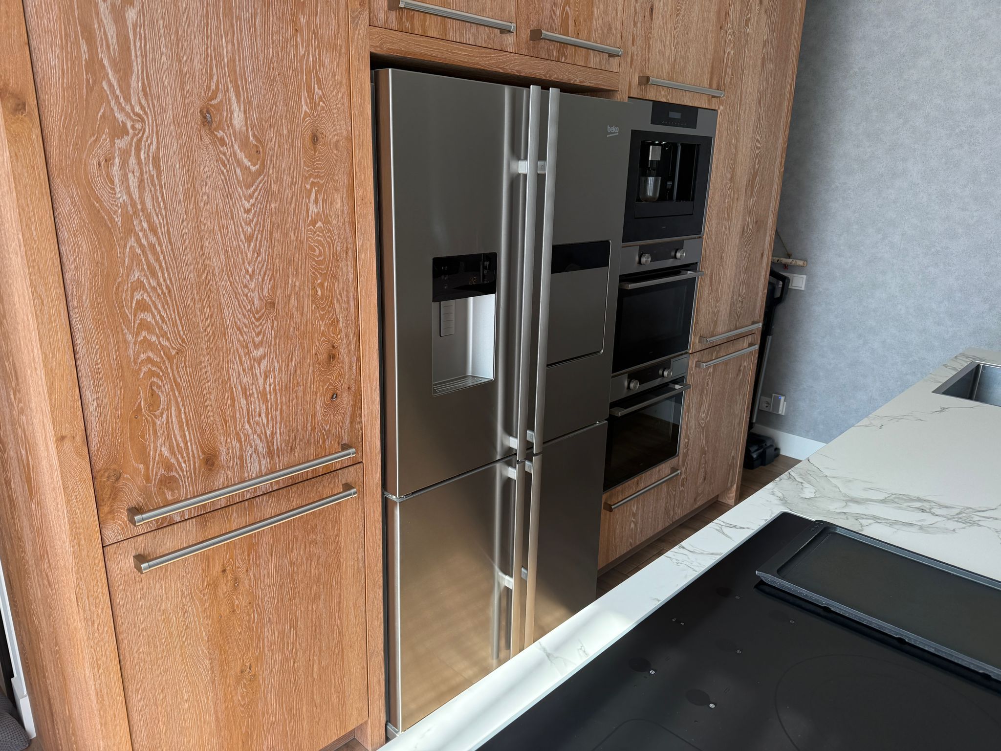 Tweedehandse keuken in houtlook met een grijs kookeiland en kunststoffen blad met grote amerikaanse koelkast, twee ovens en ingebouwd koffiezetapparaat